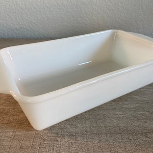 May include: White ceramic loaf pan with a smooth, glossy finish. The pan has a rectangular shape with slightly rounded edges and two small handles on the sides.