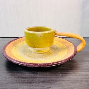 May include: A yellow and brown ceramic candle holder with a handle. The holder has a ring of brown around the top and bottom.