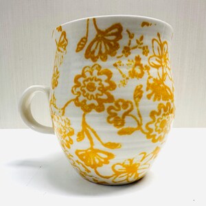 May include: White ceramic mug with a curved handle, decorated with a repeating pattern of yellow floral designs. The mug has a wide opening and a slightly textured surface, suitable for hot drinks.