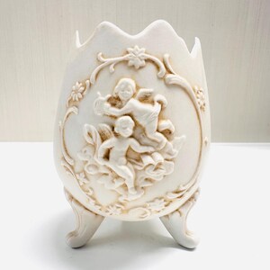 May include: Off-white, egg-shaped decorative vase with three curved feet. The vase features a relief design of cherubs and floral accents. The top edge has a scalloped design. A classic, elegant home decor item.