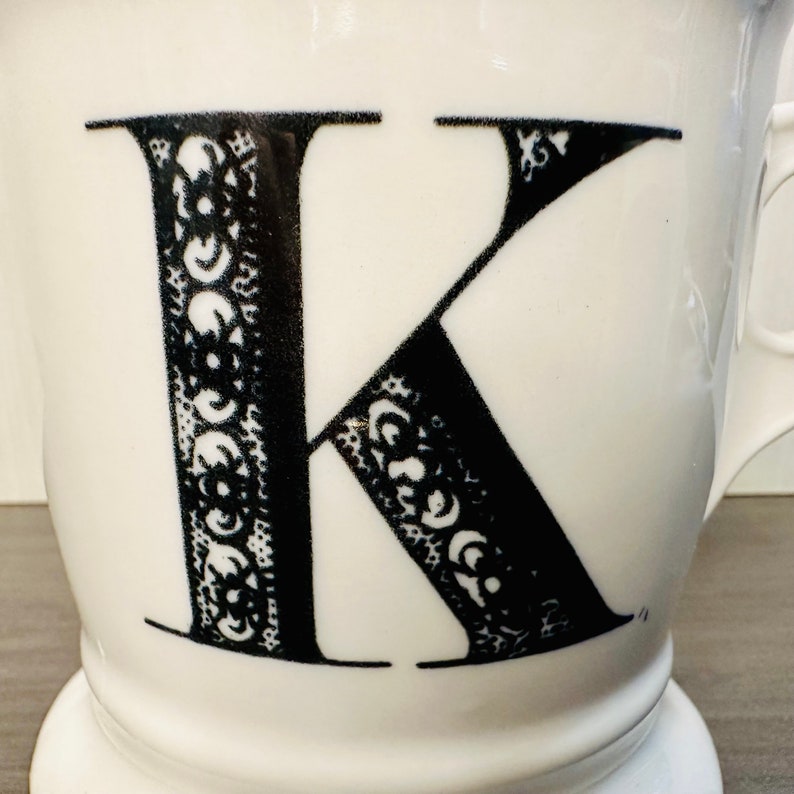May include: White ceramic mug with a large black letter "K" decorated with an ornate floral pattern. The mug has a wide base and a curved handle, ideal for tea or coffee.