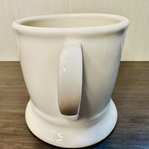 May include: Cream-coloured ceramic mug with a rounded base and a handle. The mug has a wide opening and a smooth, glossy finish. The handle is a lighter shade of cream, with a darker brown gradient at the base.