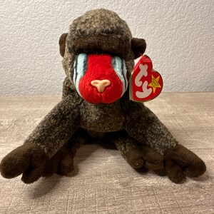 May include: A brown and gray stuffed monkey with a red face and blue stripes. The monkey has a Ty Beanie Baby tag attached to its ear. The tag is red with a white star and the words "Beanie Babies" in white.