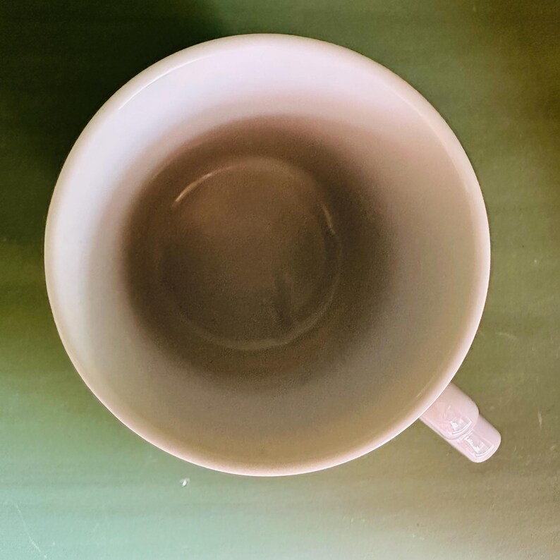 K&ouml;nnte beinhalten: Draufsicht auf eine leere, cremefarbene Keramiktasse mit Henkel. Das Innere der Tasse ist hellbraun. Die Tasse steht vor einem gr&uuml;nen Hintergrund.