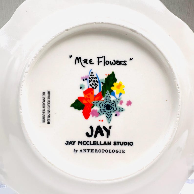 May include: White ceramic plate with a scalloped edge, featuring a colorful floral design and the text "Mae Flowers." The plate includes the text "JAY" and "JAY MCCLELLAN STUDIO by ANTHROPOLOGIE."