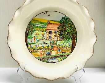 May include: A decorative ceramic plate with a scalloped edge and a painted scene of a cottage and people in a garden. The plate has a cream-colored background with a brown trim.