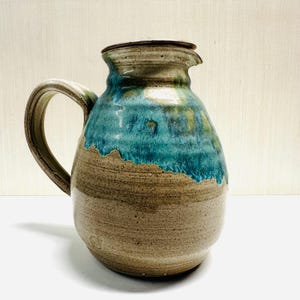 May include: A handmade ceramic pitcher with a lid. The pitcher features a two-tone design, with a tan base and a turquoise blue upper section. The handle is curved, and the pitcher has a spout for pouring. The pitcher is approximately 10 inches tall.