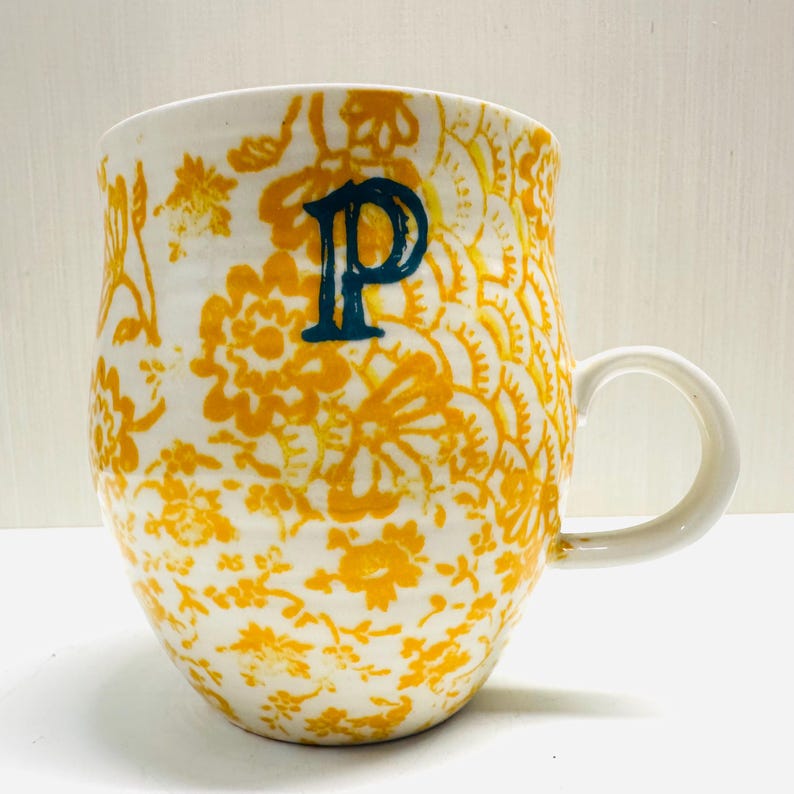 May include: White ceramic mug with a yellow floral pattern and a teal letter "P". The mug has a curved shape and a white handle. The floral design covers the entire surface of the mug.