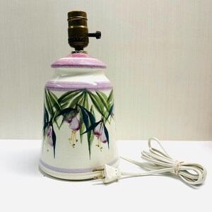 May include: A ceramic table lamp with a white base, adorned with a floral design featuring purple and green accents. The lamp has a brass-colored socket and a white power cord. The lamp's design includes a purple band around the top and bottom.