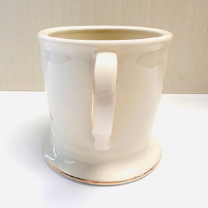 Puede incluir: Una taza de cer&aacute;mica blanca con dos asas y un borde dorado alrededor de la parte superior e inferior. La taza est&aacute; vac&iacute;a.