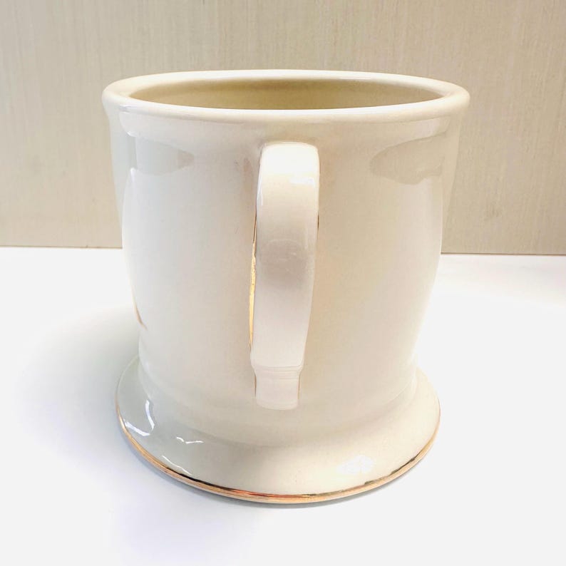 Puede incluir: Una taza de cer&aacute;mica blanca con dos asas y un borde dorado alrededor de la parte superior e inferior. La taza est&aacute; vac&iacute;a.