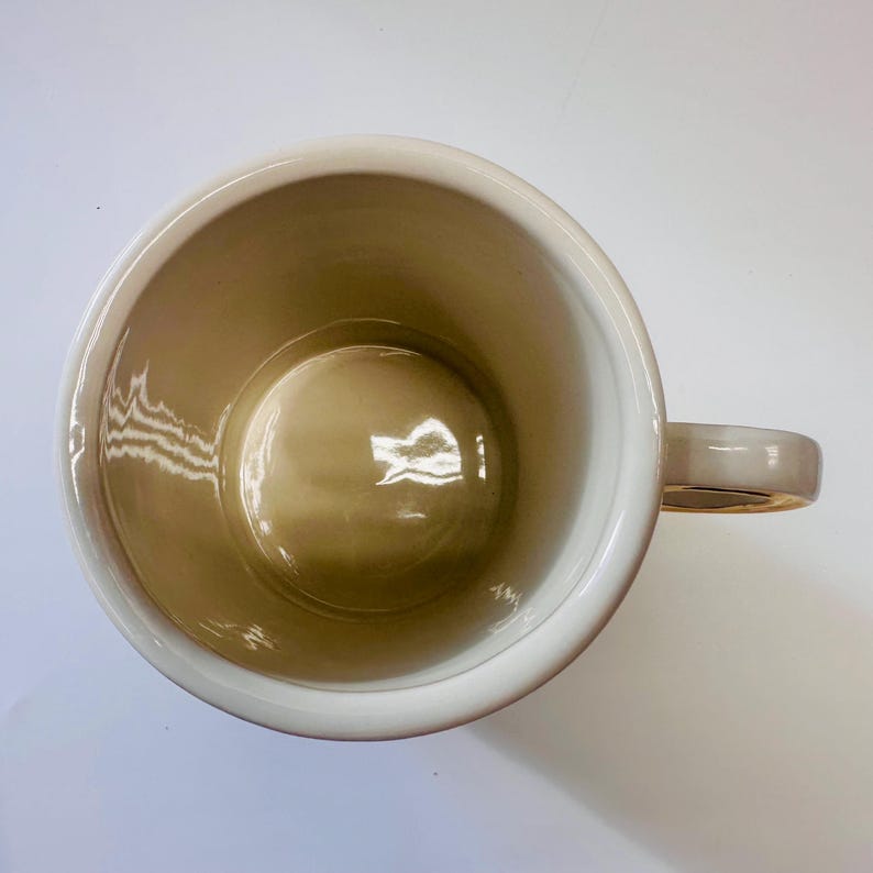 Puede incluir: Una taza de cer&aacute;mica blanca con un borde y un asa dorados. El interior de la taza es de color beige claro.