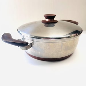 May include: A stainless steel cooking pot with a lid and two handles. The pot has a brown base and brown handles. The lid has a brown knob. The pot is sitting on a white surface.
