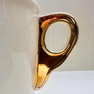 Puede incluir: Una taza de cer&aacute;mica blanca con un asa de color dorado. El asa tiene un dise&ntilde;o decorativo y es brillante.