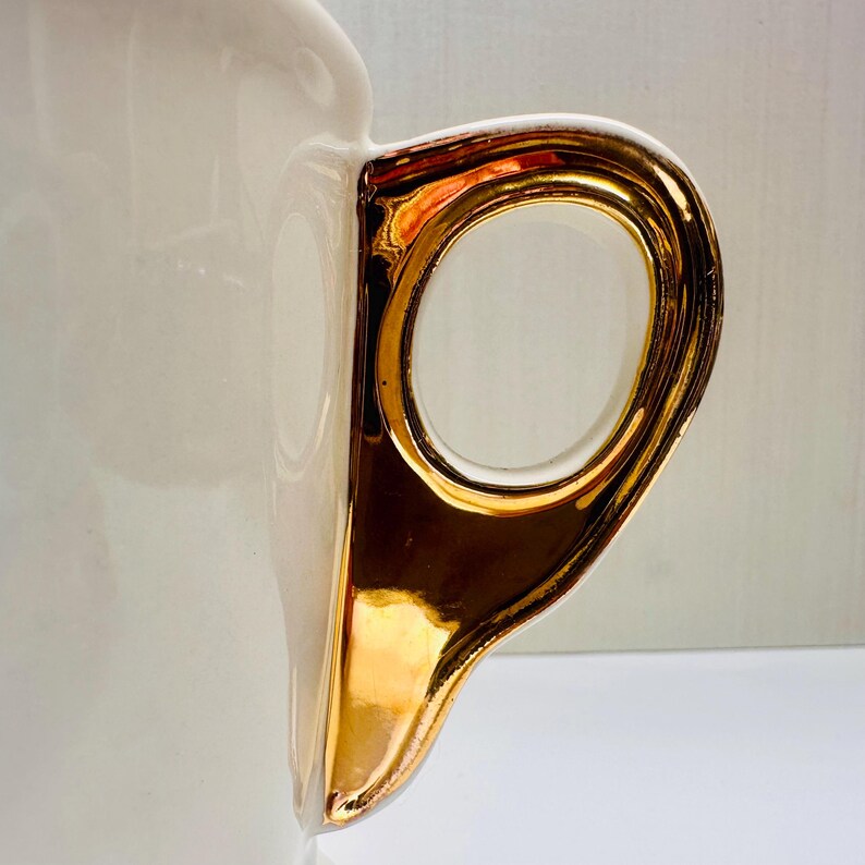 Puede incluir: Una taza de cer&aacute;mica blanca con un asa de color dorado. El asa tiene un dise&ntilde;o decorativo y es brillante.