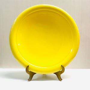 May include: A bright yellow ceramic plate with a glossy finish. The plate is round and sits on a decorative brass stand. The plate's vibrant color makes it a striking decorative piece.