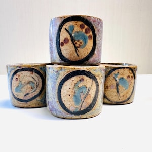 May include: A stack of four handmade ceramic cups. Each cup features a unique design within a black circle, with blue and brown abstract shapes. The cups have a speckled, earthy color palette with hints of purple.
