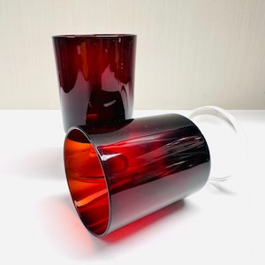 May include: Two ruby red drinking glasses. One glass is upright, and the other is on its side. The glasses are made of a transparent material, and the interior of the glasses is visible. The glasses are on a white surface.