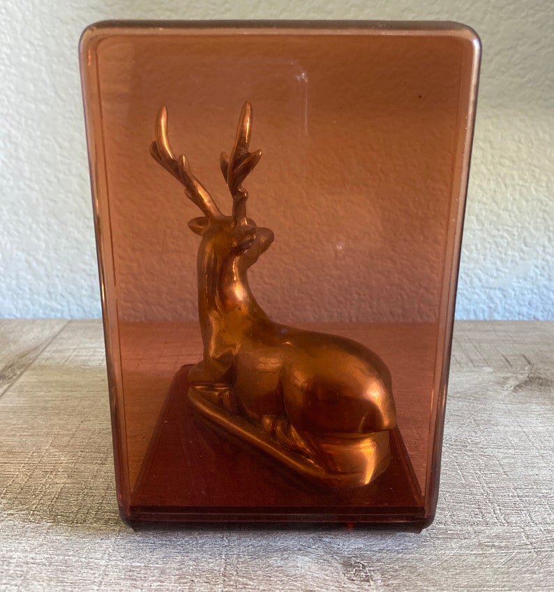 Puede incluir: Una figurita de ciervo de color cobre con cuernos se encuentra dentro de una caja de vidrio transparente rectangular de color &aacute;mbar.
