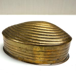 May include: An oval-shaped brass box with a hinged lid. The lid features a series of concentric, raised ridges that mimic the appearance of a seashell. The sides of the box are smooth with horizontal engraved lines encircling the entire base.