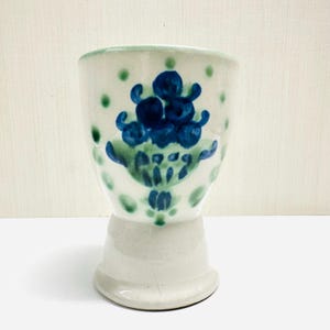 May include: A white ceramic egg cup with a flared base and a blue and green floral design. The cup features a hand-painted design of blue flowers and green leaves, with green polka dots around the rim.