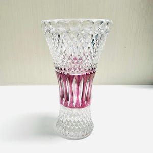 May include: A clear glass vase with a diamond-cut pattern. The vase has a flared top and a base with a similar design. A band of magenta-colored glass accents the center of the vase, adding a pop of color.