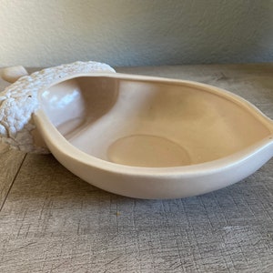 May include: A light beige ceramic acorn-shaped bowl. The bowl has a smooth, glossy interior and a textured, bumpy exterior on the acorn cap. The bowl is designed with a leaf-like shape.