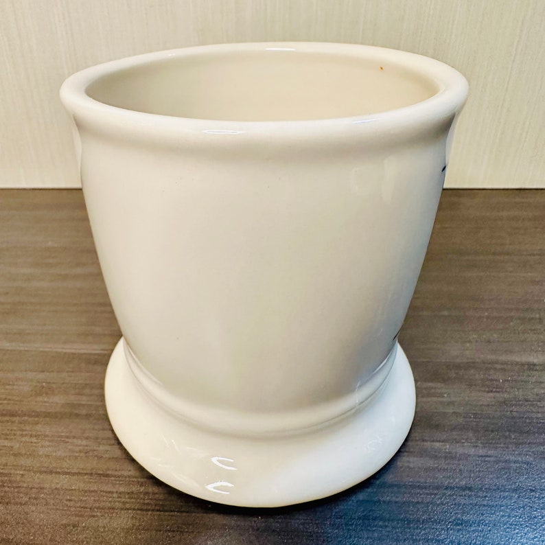 May include: A white ceramic shaving mug with a flared top and a pedestal base. The mug has a smooth, glossy finish and a simple design. The mug is empty and ready to be filled with shaving cream.
