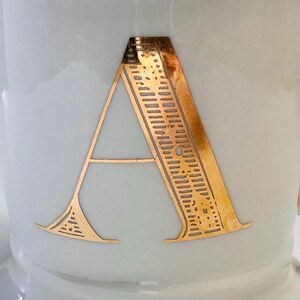 Puede incluir: Una taza de cer&aacute;mica blanca con una letra "A" dorada en la parte delantera. La letra est&aacute; rodeada por un patr&oacute;n decorativo.
