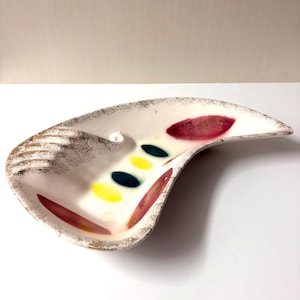 May include: A crescent-shaped ceramic dish with an off-white base, featuring a gold-flecked rim and decorative accents. The interior displays a series of colorful painted ovals in yellow, teal, and deep red, arranged in a linear pattern.