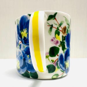 May include: White ceramic mug with a floral design featuring large blue flowers, green leaves, and accents of pink and yellow. The handle is white with a yellow stripe down the center. The mug is a perfect gift for a coffee or tea lover.