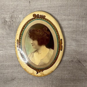 May include: An oval-shaped, cream-colored compact mirror with a gold and blue decorative border. The mirror features a vintage photograph of a woman with dark hair, wearing a light-colored dress.
