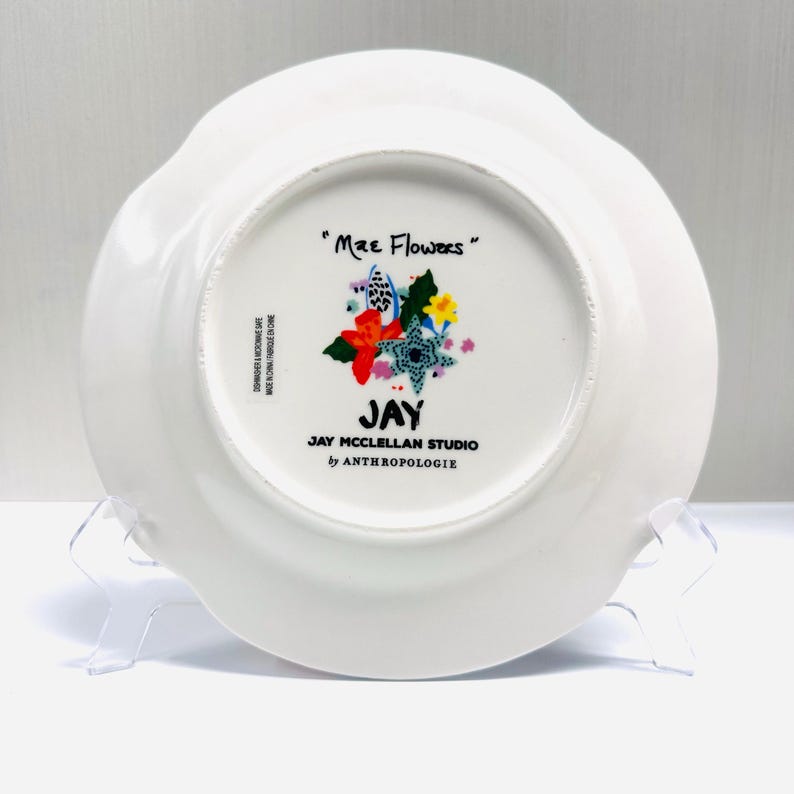 May include: White ceramic plate with a scalloped edge. The bottom of the plate features a floral design with the text "Mae Flowers" and "JAY JAY MCCLELLAN STUDIO by ANTHROPOLOGIE".