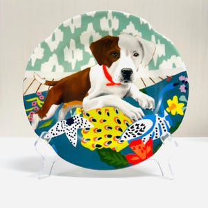 May include: A decorative plate featuring a dog illustration. The dog has brown and white fur and a red collar. The plate has a teal background with floral and abstract patterns in yellow, blue, and red. The plate is displayed on a clear stand.