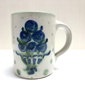 May include: A ceramic mug with a speckled off-white finish featuring a hand-painted blue and green cluster of berries or fruit in a bowl design. The mug has a curved handle on the right side and scattered light green dots across the surface.