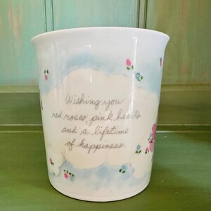 K&ouml;nnte beinhalten: Wei&szlig;er Keramikbecher mit hellblauem und wei&szlig;em Wolkenmuster, verziert mit kleinen rosa und blauen Blumenakzenten. Der Becher tr&auml;gt den handgeschriebenen Text: "Wishing you red roses, pink hearts and a lifetime of happiness."