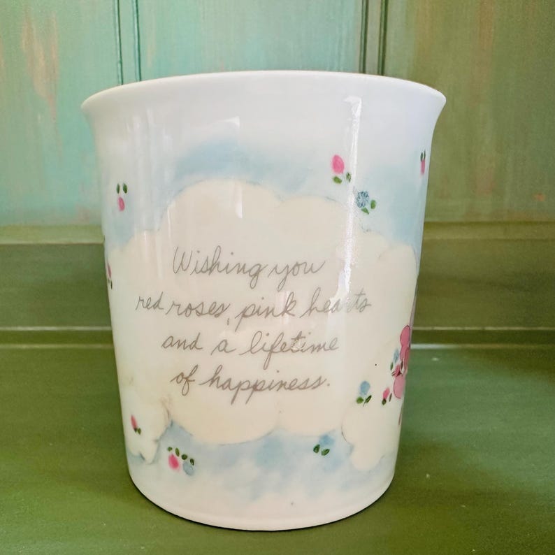 K&ouml;nnte beinhalten: Wei&szlig;er Keramikbecher mit hellblauem und wei&szlig;em Wolkenmuster, verziert mit kleinen rosa und blauen Blumenakzenten. Der Becher tr&auml;gt den handgeschriebenen Text: "Wishing you red roses, pink hearts and a lifetime of happiness."