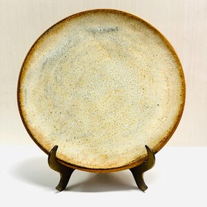 May include: A round ceramic plate with a speckled, off-white center and a brown rim. The plate is displayed on a decorative bronze stand. The plate has a rustic, handmade appearance.