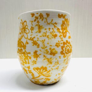May include: White ceramic vase featuring a yellow floral pattern. The vase has a rounded shape and a wide opening. The yellow floral design covers the entire surface, creating a decorative look. Suitable for flowers or as a decorative item. The vase measures approximately 15 cm in height.