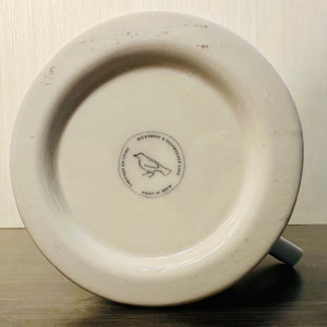 May include: Underside of a white ceramic mug with a grey handle. The base has a circular emblem with a bird illustration and the text "MICROWAVE & DISHWASHER SAFE", "FABRIQU&Eacute; EN CHINE", and "MADE IN CHINA".