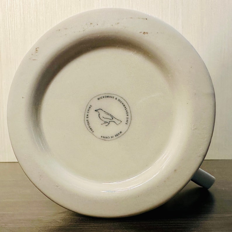 May include: Underside of a white ceramic mug with a grey handle. The base has a circular emblem with a bird illustration and the text "MICROWAVE & DISHWASHER SAFE", "FABRIQU&Eacute; EN CHINE", and "MADE IN CHINA".