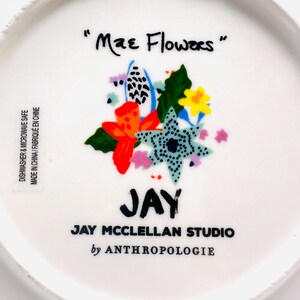 May include: White ceramic plate with the text "Mae Flowers" and a colorful floral design. The plate features the name "JAY" and "JAY MCCLELLAN STUDIO by ANTHROPOLOGIE". Dishwasher and microwave safe.