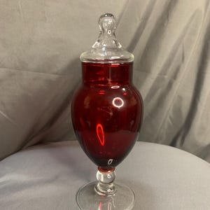 May include: A red glass candy jar with a clear glass lid and a clear glass base. The jar has a round body and a wide mouth.