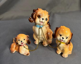 May include: Three ceramic dog figurines in shades of brown and tan, each with a different colored collar. The dogs are connected by a gold-colored chain leash. The dogs have black eyes and pink tongues.