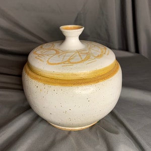 May include: White ceramic jar with a lid, featuring a brown geometric design. The jar has a speckled glaze and a rounded shape.