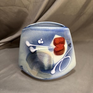 May include: A ceramic vase with a wide, tapered shape and a blue gradient finish. The surface features abstract painted details including a dark red rectangular shape, white linear accents, and a circular dark blue patch with a glossy glaze.