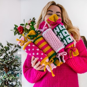May include: Four knitted Christmas cracker decorations in pink, yellow, green, and white with a heart pattern. The crackers are tied with a yellow ribbon and have a green bow.