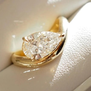 May include: A gold ring featuring a pear-cut diamond, presented in a white box. The ring's band elegantly curves around the diamond. The diamond is clear and reflects light, creating a sparkling effect.