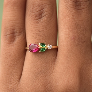 Peut inclure: Une bague en or avec une pierre ovale rouge, deux pierres ovales vertes et une petite pierre ronde blanche. La bague est portée sur un doigt.