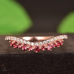 May include: A delicate, curved wedding band with a double row of sparkling stones. The bottom row features red gemstones, while the top row is set with clear stones.